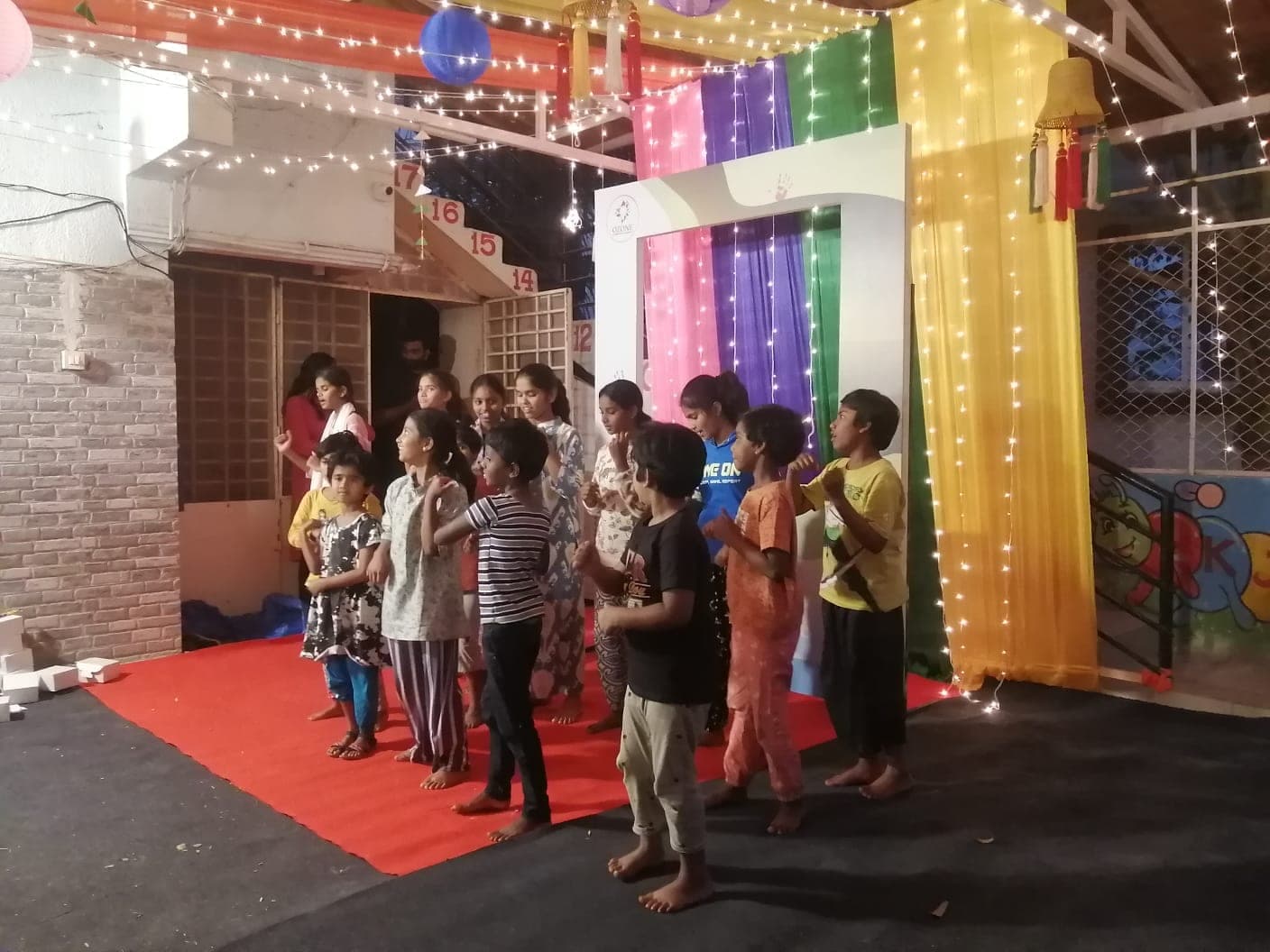Children performing on a red carpet stage under strings of fairy lights and coloured drapes.