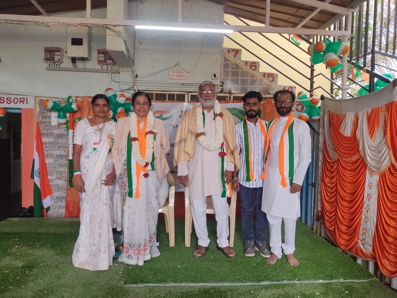 Five adults standing together for a photograph in front of Indian flags and festive decorations.